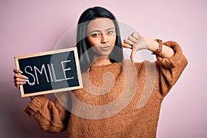 Young chinese woman holding blackboard with smile word over isolated pink background with angry face, negative sign showing