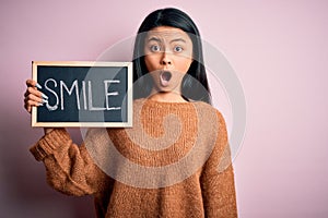 Young chinese woman holding blackboard with smile word over isolated pink background scared in shock with a surprise face, afraid