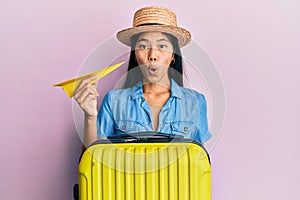 Young chinese woman holding cabin bag and paper plane scared and amazed with open mouth for surprise, disbelief face