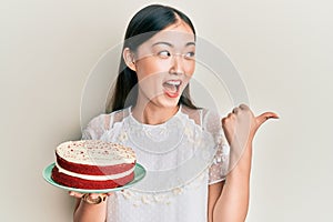 Young chinese woman holding carrot cake pointing thumb up to the side smiling happy with open mouth