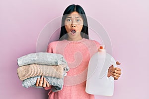 Young chinese woman holding detergent bottle and clothes afraid and shocked with surprise and amazed expression, fear and excited