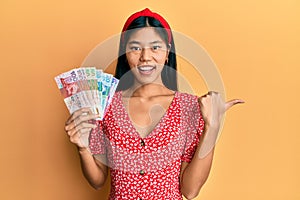 Young chinese woman holding hong kong dollars banknotes pointing thumb up to the side smiling happy with open mouth