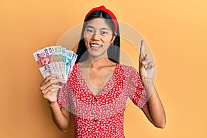 Young chinese woman holding hong kong dollars banknotes smiling with an idea or question pointing finger with happy face, number