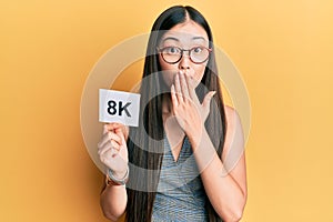 Young chinese woman holding 8k resolution technology paper covering mouth with hand, shocked and afraid for mistake photo