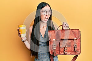 Young chinese woman holding leather bag and drinking a take away coffee angry and mad screaming frustrated and furious, shouting