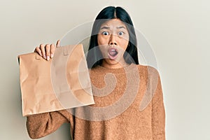 Young chinese woman holding take away paper bag scared and amazed with open mouth for surprise, disbelief face