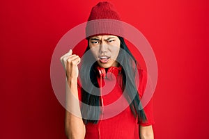 Young chinese woman listening to music using headphones angry and mad raising fist frustrated and furious while shouting with