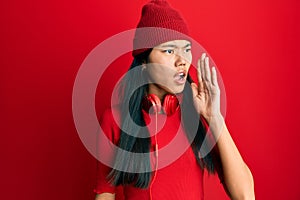 Young chinese woman listening to music using headphones shouting and screaming loud to side with hand on mouth