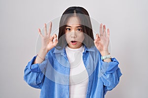 Young chinese woman standing over white background looking surprised and shocked doing ok approval symbol with fingers