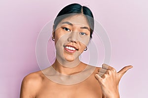 Young chinese woman standing topless showing skin smiling with happy face looking and pointing to the side with thumb up