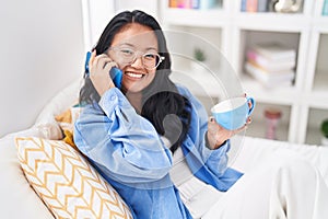 Young chinese woman talking on smartphone drinking coffee at bedroom