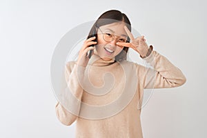 Young chinese woman talking on the smartphone over isolated white background Doing peace symbol with fingers over face, smiling