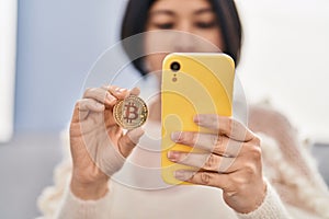Young chinese woman using smartphone holding bitcoin at home