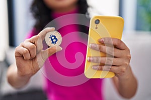 Young chinese woman using smartphone holding bitcoin at home