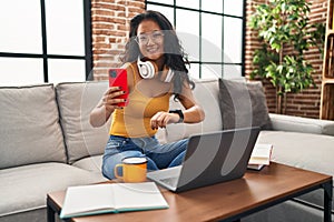 Young chinese woman using smartphone looking watch sitting on sofa at home