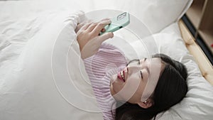 Young chinese woman using smartphone lying on bed at bedroom