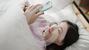 Young chinese woman using smartphone lying on bed at bedroom