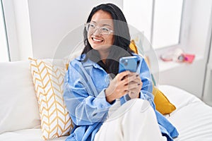 Young chinese woman using smartphone sitting on bed at bedroom