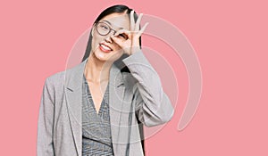 Young chinese woman wearing business clothes smiling happy doing ok sign with hand on eye looking through fingers