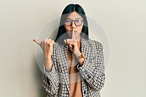 Young chinese woman wearing business style and glasses asking to be quiet with finger on lips pointing with hand to the side