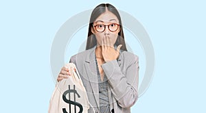 Young chinese woman wearing business suit holding dollars bag covering mouth with hand, shocked and afraid for mistake