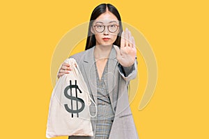 Young chinese woman wearing business suit holding dollars bag with open hand doing stop sign with serious and confident