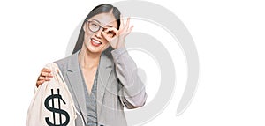 Young chinese woman wearing business suit holding dollars bag smiling happy doing ok sign with hand on eye looking through fingers