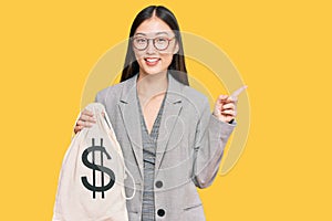 Young chinese woman wearing business suit holding dollars bag smiling happy pointing with hand and finger to the side