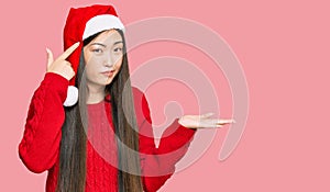 Young chinese woman wearing christmas hat confused and annoyed with open palm showing copy space and pointing finger to forehead