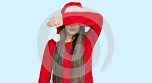 Young chinese woman wearing christmas hat covering eyes with arm, looking serious and sad