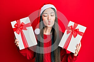 Young chinese woman wearing christmas hat and holding gifts making fish face with mouth and squinting eyes, crazy and comical