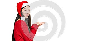 Young chinese woman wearing christmas hat pointing aside with hands open palms showing copy space, presenting advertisement