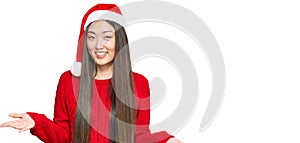 Young chinese woman wearing christmas hat smiling cheerful with open arms as friendly welcome, positive and confident greetings