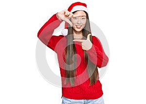 Young chinese woman wearing christmas hat smiling making frame with hands and fingers with happy face