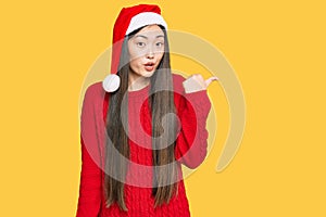 Young chinese woman wearing christmas hat surprised pointing with hand finger to the side, open mouth amazed expression