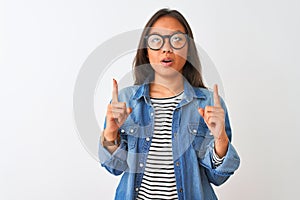 Young chinese woman wearing denim shirt and glasses over isolated white background amazed and surprised looking up and pointing