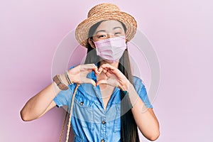 Young chinese woman wearing medical mask on summer smiling in love doing heart symbol shape with hands