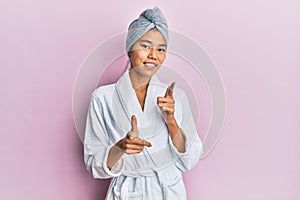 Young chinese woman wearing shower towel cap and bathrobe pointing fingers to camera with happy and funny face