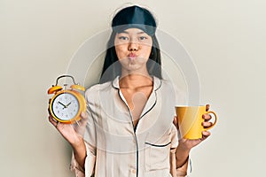 Young chinese woman wearing sleep mask and robe holding alarm clock and cup of coffee puffing cheeks with funny face