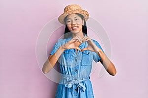 Young chinese woman wearing summer hat smiling in love doing heart symbol shape with hands