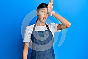 Young chinese woman wearing waiter apron surprised with hand on head for mistake, remember error