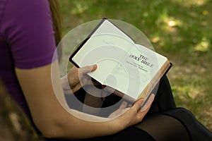 Young Christian woman holding and reading an open Holy Bible Book in peace in nature