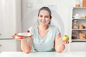 Making Right Choice. Chubby girl sitting at kitchen choosing between plate with desserts and apple looking camera