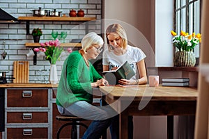 Young church volunteer reading Bible with pensioner at home kitchen