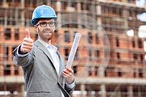 Young civil engineer or businessman standing in front of a construction site and showing thumbs up