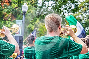 Young clean cut men in sweaty green t shirts play trumpets in parade with bokeh trees and American flag in background - back view