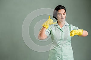 Young cleaning woman wearing a green shirt and yellow gloves showing it`s time gesture