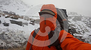 Young climber man with backpack, red coat and cap taking a selfie while climbing a snowy mountain