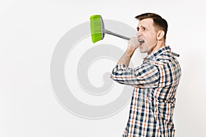 Young combative angry housekeeper man in checkered shirt holding and sweeping with green broom isolated on white