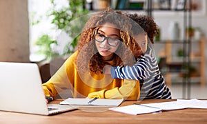 Young concentrated afro american woman mother working online on laptop with kid son at home